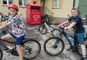 Chłopiec i dziewczyna siedzący na rowerach. Za nim stoją rowery. W tle żólty budynek i czerwony pojemnik na odzież.