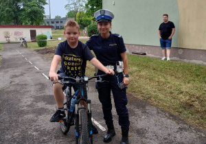 Chłopiec siedzi na rowerze. Przy nim stoi pani policjantka. W tle żółty budynek i stojący przed nim ubrany na czarno mężczyzna.