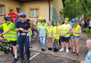 Na pierwszym planie stoi policjantka. Obok niej na rowerze w kamizelce odblaskowej i kasku siedzi chłopiec. Za nimi stoi duża grupa dzieci w kamizelkach odblaskowych. W tle żółty budynek.