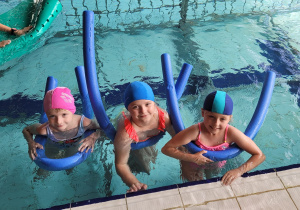 Trzy dziewczynki w wodzie pozują do zdjęcia. Dziewczynki pływaja na piankach rurowych wygiętych w literę U.