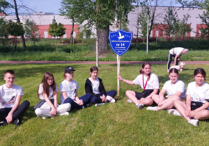 Na trawie w szeregu siedzi grupa biegaczy w białych koszulkach. Jedna z dziewczyn trzyma tarczę szkoły . W tle drzewa i budynek.