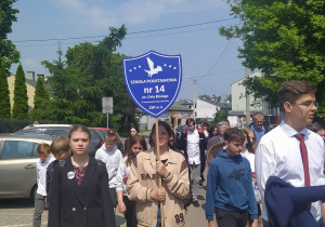 Duża grupa uczniów idzie ulicą. Na pierwszym planie uczniowie z tarczą. Po lewej stronie widoczne samochody na parkingu.