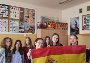 Wnętrze klasy. Dziewczęta stoją w szeregu. W rekach trzymają dużą flagę Hiszpani. W tle sciana z gazetkami szkolnymi.