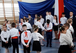 Uczniowie w galowych strojach tańczą. Za nimi niebieskie tło i flaga Polski.