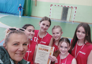 Hala sportowa. Pięć dziewcząt w czerwonych strojach sportowych i trenerka pozują do zdjęcia z medalami i dyplomem.