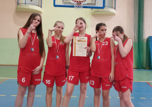 Hala sportowa. Pięć dziewcząt w czerwonych strojach sportowych pozuje do zdjęcia z medalami i dyplomem.