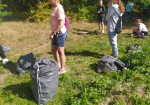 Grupa uczniów stoi na tle lasu. Przed uczniami stoją szare worki ze śmieciami