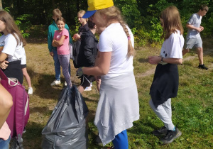 Grupa uczniów stoi na tle lasu. Przed uczniami stoją szare worki ze śmieciami