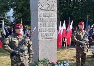 Żołnierze pełnią wartę przy pomniku Sybiraków. Przed pomnikiem leżą białe i czerwone kwiaty. W tle drzewa i Poczty sztandarowe.
