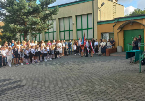 Plac przed szkołą. Na placu stoją uczniowie w dwuszeregu. Za nimi stoją rodzice. Po prawej stronie przy stoliku stoi kobieta. W tle budynek szkolny.