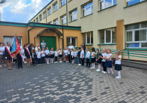 Plac szkolny. Na placu w szeregu stoją dzieci ubrane na galowo. Po lewej stronie stoi chłopiec trzymający sztandar szkoły. Po jego prawej i lewej stronie stoją uczennice. W tle budynek szkolny.