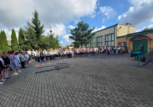 Plac przed szkołą. Na placu stoją uczniowie w dwuszeregu. Za nimi stoją rodzice. Po prawej stronie przy stoliku stoi kobieta. W tle budynek szkolny i drzewa iglaste.