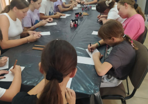 Duży stół przykryty czarna folią. Wokół stołu siedzą dziewczęta i rysują na kartkach. Na stole stoją lakiery do paznokci.