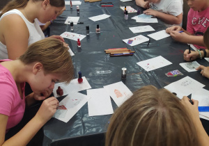Duży stół przykryty czarna folią. Wokół stołu siedzą dziewczęta i malują kamieniczki wygrawerowane na szkle. Na stole stoją lakiery do paznokci, leżą ołówki i mazaki.