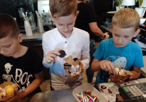 Wnętrze kawiarni. Trzech chłopców stoi przy bufecie. Uczniowie trzymają w ręku ogromy rożek z deserem lodowym. Na bufecie przed nimi leżą plastikowe łyżeczki. W tle wnętrze zaplecze kuchenne.