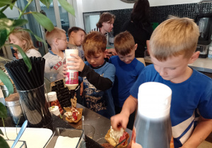Wnętrze kawiarni. Chłopiec w niebieskiej bluzce stoi przy bufecie. Uczeń ma przed sobą ogromy rożek z deserem lodowym. Na bufecie przed nimi stoją butelki. W tle grupa uczniów i wnętrze zaplecza kuchennego.