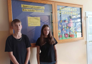 Chłopiec i dziewczyna w czarnych koszulkach stoją na tle ściany. Na ścianie wisi gablota z informacjami Samorządu Uczniowskiego.