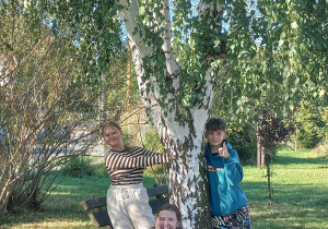 Ogromna brzoza. Przed brzozą kucnęła dziewczyna. Dwie dziewczyny ustawione po prawej i po lewej stronie drzewa. Jedna z nich przytula się do brzozy. Druga dziewczyna obejmując brzozę lekko się odchyla od drzewa. W tle krzewy.