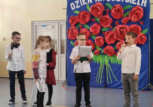 Sala gimnastyczna. Grupa dzieci ubranych na galowo stoi na tle dekoracji. Dekoracja to czerwone róże z papieru przypięte na niebieskim tle.