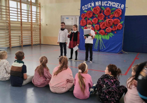 Sala gimnastyczna. Przed trójką uczniów siedzi na podłodze rząd dzieci. W tle dekoracja z różami .