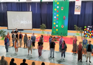 Na pierwszym planie w dwuch szeregach stoi duża grupa uczniów ubrana w kolorowe koszule z wiankami na szyjach. W tle ekran i wisząca w pionie zielona tkaninatkanina