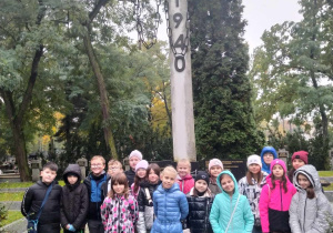 Dzieci stoja przed dużym betonowym krzyżem z napisem Katyń. Na krzyżu wisi łańcuch.
