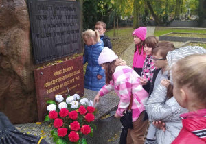 Grupa dzieci stoi przed wielkim kamieniem. Na kamieniu umocowana jest płyta z napisaem " pamięci żołnierzy poległych..." przed płyta lezy wieniec z białych i czerwonych kwiatów.