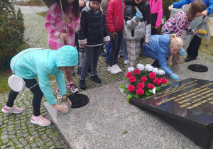 Grupa dzieci stoi przed czarna płytą leżącą na szarym postumencie. przed płyta lezy wieniec z białych i czerwonych kwiatów. Jedno z dzieci stawia przed płytą znicz.