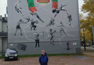 Chłopeic stoi na tle ściany bocznej kamienicy. Na ścianie znajduje się ogromny mural. Mural poświęcony jest 100 -leciu klubu sportowego Lechii.