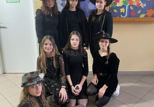 Korytarz szkolny. Siedem dziewcząt ubranych na czarno pozuje do zdjęcia. Trzy z nich stoją .Trzy kucnęły a jedna usiadła na podłodze bokiem do reszty dziewcząt.