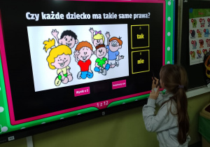 Tablica interaktywna z wyswietlonym obrazkiem dzieci. Przed tablicą stoi uczennica.