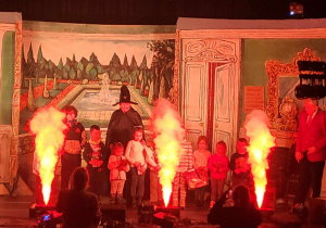 Scena. Na scenie grupa aktorów i uczniów stoi na tle pałacowego wnętrza. Przed nimi palą się trzy duże race.