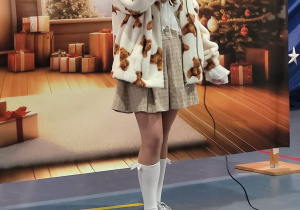 Uczennica ubrana w białe futerko z brązowymi misiami stoi z mikrofonem w ręku. Za nią ekran z wnętrzem mieszkania w świątecznym wystroju.