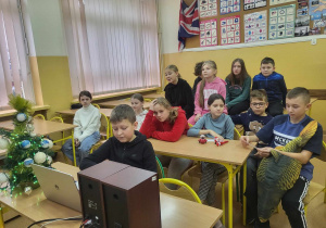 Klasa szkolna. Na pierwszym planie przed laptopem siedzi chłopiec. Obok laptopa stoi mała choinka. Za chłopcem w ławkach siedzą uczniowie. W tle na ścianie wiszą tablice dydaktyczne.