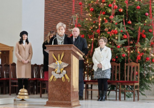 Wnętrze kościoła. Na pierwszym planie stoi mównica a przy niej kobieta. Za nią stoją trzy kobiety i mężczyzna. W tle ogroma choinka ubrana w czerwone ozdoby.