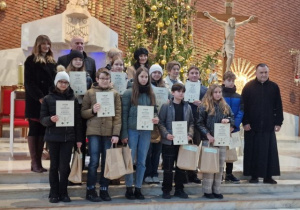 Wnętrze kościoła. Przy ołtarzu stoi duża grupa uczniów z dyplomami w rękach. Po prawej stronie stoi ksiądz. Za dziećmi stoją trzy kobiety i mężczyzna. W tle widoczny ołtarz i choinka ubrana w złote ozdoby.