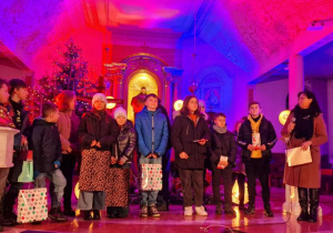 Wnętrze kościoła podświetlone czerwonym i niebieskim światłem. Przed ołtarzem stoi grupa kilkunastu dzieci. Po prawej stronie stoi kobieta z mikrofonem. W tle ołtarz.