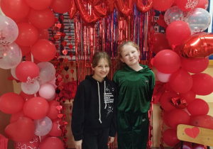 Dwie dziewczynki stoją na tle walentynkowej ścianki. Walentynkowa ścianka to łuch z dużej ilości czerwonych i białych balonów. Łuk wypełniony jest czerwoną lametą.