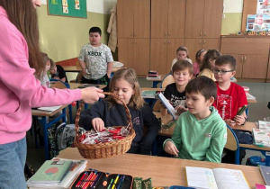 Wnętrze klasy. Dziewczyna z koszykiem rozdaje uczniom lizaki w kształcie serduszek.