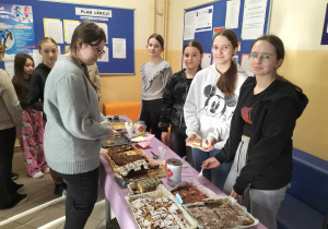 Korytarz szkolny. Na środku zdjęcia znajduję się stół z różnymi ciastami. Na stole stoją puszki z logo Caritas. Po prawej stronie stołu stoją cztery uczennice sprzedające ciasta. Po lewej stronie stoją kupujący ciasta.