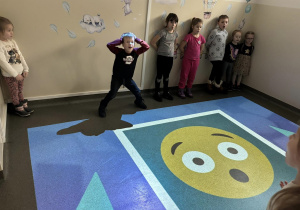 Wnętrze pracowni. Grupka dzieci stoi oparta o ścianę. Na podłodze wyświetla się obraz z emotikonką.