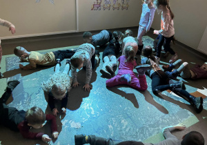 Wnętrze pracowni.Grupka dzieci lerzy swobodnie na brzuchach . Dwie dziewczynki stoją. Na podłodze wyświetla się obraz wody.