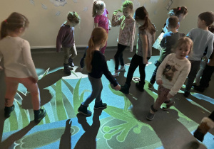 Wnętrze pracowni.Grupka dzieci stoi na oświetlonej podłodze. Na podłodze wyświetla się obraz przedstawiający staw z żabkami.