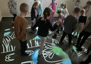 Wnętrze pracowni.Grupka dzieci stoi na oświetlonej podłodze. Na podłodze wyświetla się obraz przedstawiający staw z żabkami.