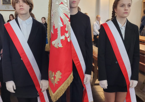 Poczet sztandarowy. Chłopiec ubrany w czarny garnitur trzyma sztandar z Orłem. Po prawej i lewej stronie chłopca stoją dwie uczennice ubrane na czarno. Wszyscy mają biało czerwone szarfy .