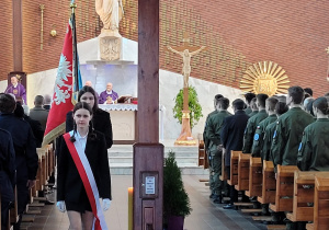 Wnętrze kościoła z widokiem na ołtarz. Po środku zdjęcia widać duży drewniany krzyż. Po lewej stronie krzyża idzie w kolumnie tyłem do ołtarza poczet sztandarowy. Na przodzie kolumny widzać dziewczynę ubraną na czarno z szarfą na klatce piersiowej. Za nią idzie chłopiec ze sztandarem. Po bokach widać ławki z uczestnikami uroczystości.