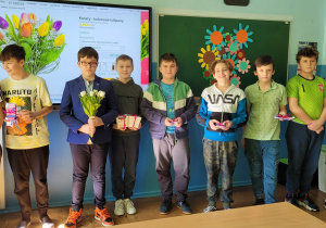 Wnętrze klasy. Grupa chłopców stoi przy ścianie. Chłopcy trzymają w rekach kwiaty i słodkości. W tle tablica interaktywna z wyświetlonym bukietem . Obok tablicy wisi tablica szkolny z przypiętymi kwiatami z papieru.
