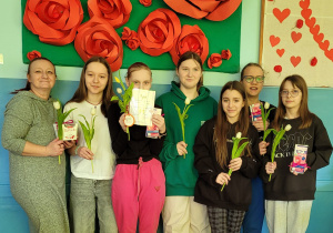 Wnętrze klasy. Kobieta i sześć dziewcząt stoją pod ścianą. Dziewczęta trzymają tulipany.Na ścianie wisi zielona tablica z czerwonymi różami z papieru.