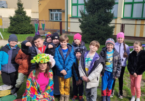 Przed szkołą. Grupa dzieci ubranych w kurtki stoi przed szkołą. Przed dziećmi przykucnęła kobieta ubrana w kapelusz z kwiatami , kolorową chustę i spódnicę. W tle plac zabaw i szkoła.