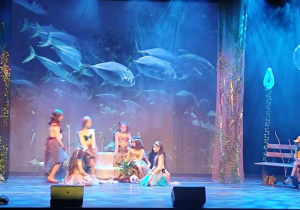 Scena z wysietlonym dnem morza. Światło na scenie w różnych odcieniach niebieskiego. Na środku sceny siedzi grupa dziewcząt.